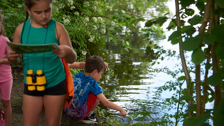 A crouching boy looks into a pond while a standing girl reads a map.